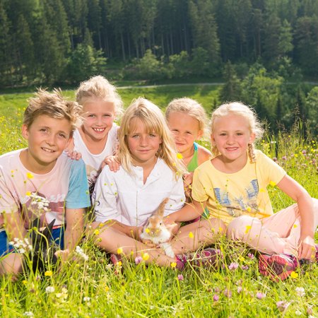 Children sit together in a flower meadow, cuddling a rabbit in the sunshine.
